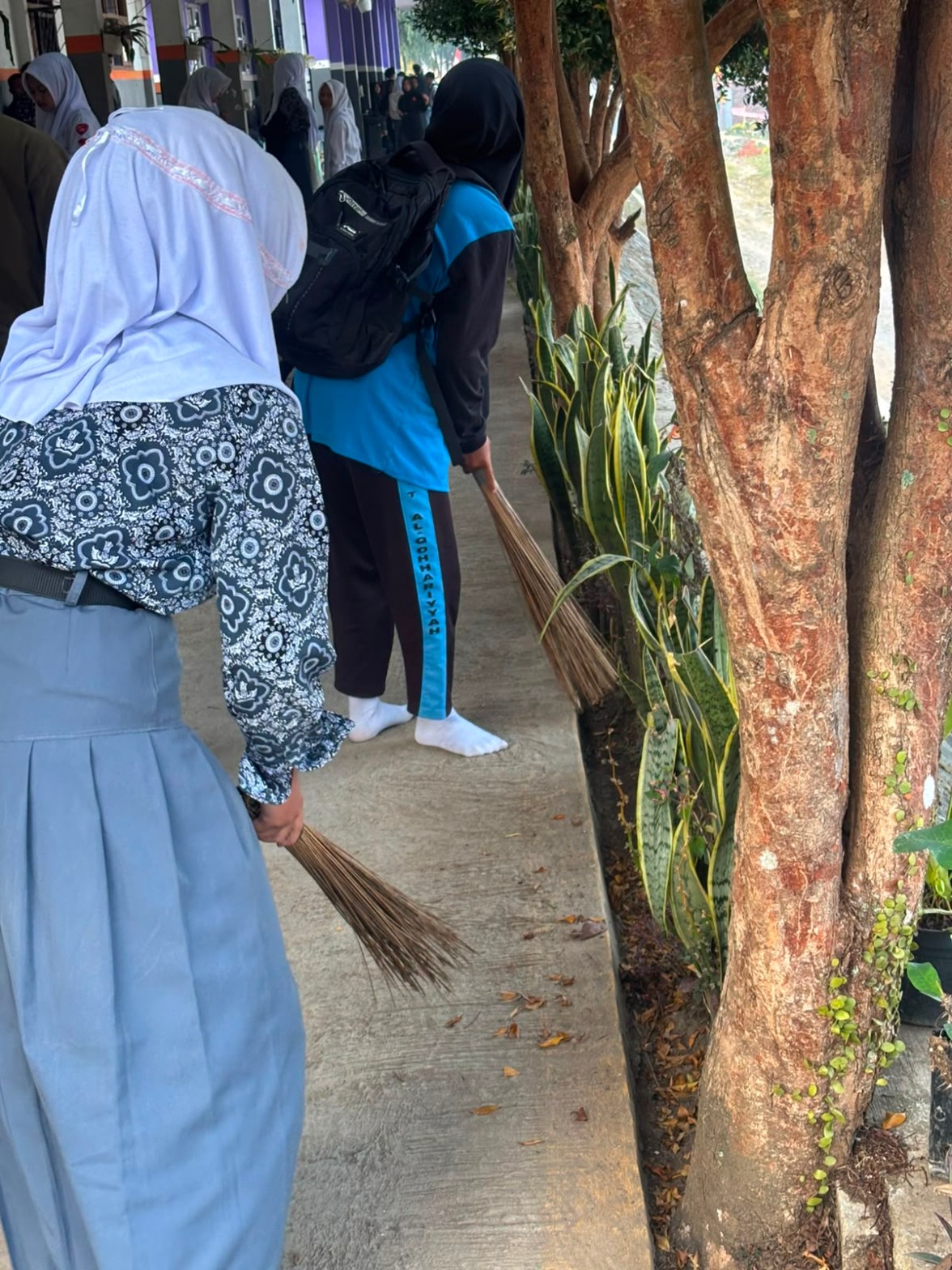 Close-up siswi menyapu daun kering di antara pohon dan tanaman sansevieria di koridor sekolah