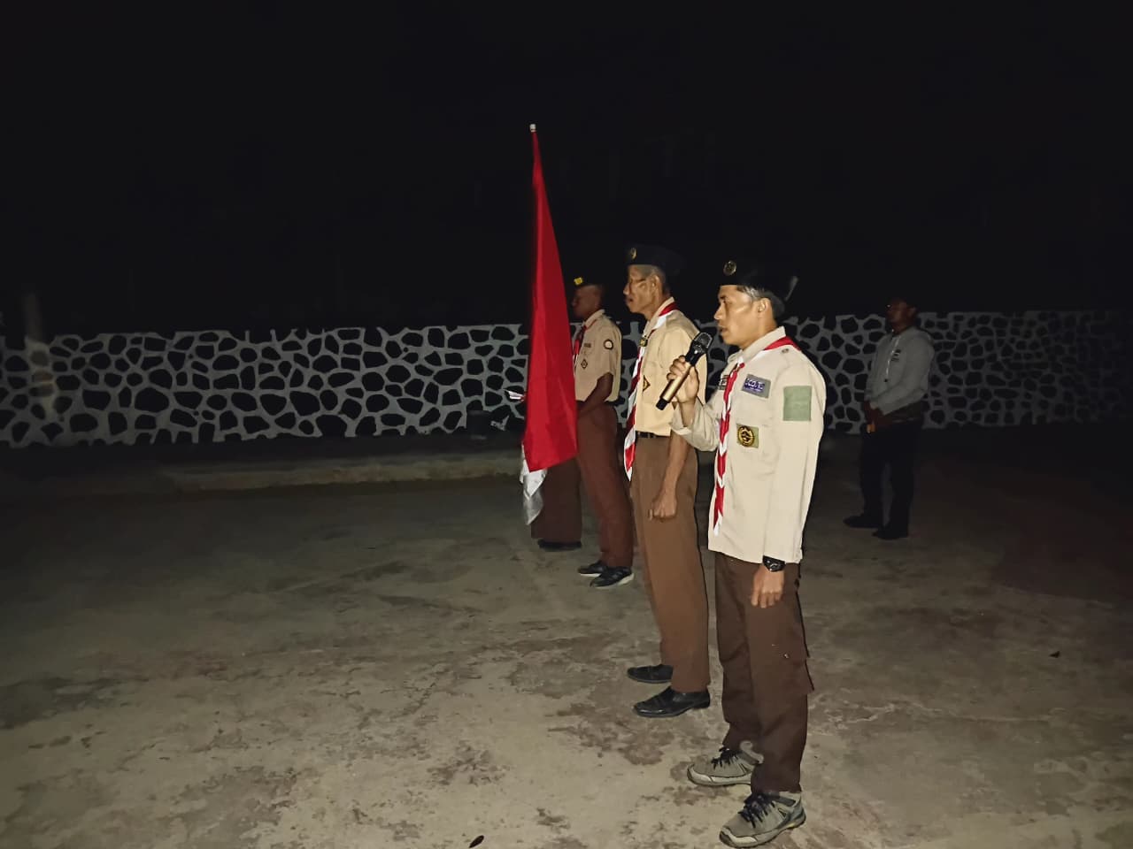 Pembina upacara menyampaikan amanat menggunakan pengeras suara pada apel malam perkemahan HUT Pramuka dengan bendera Merah Putih di sampingnya