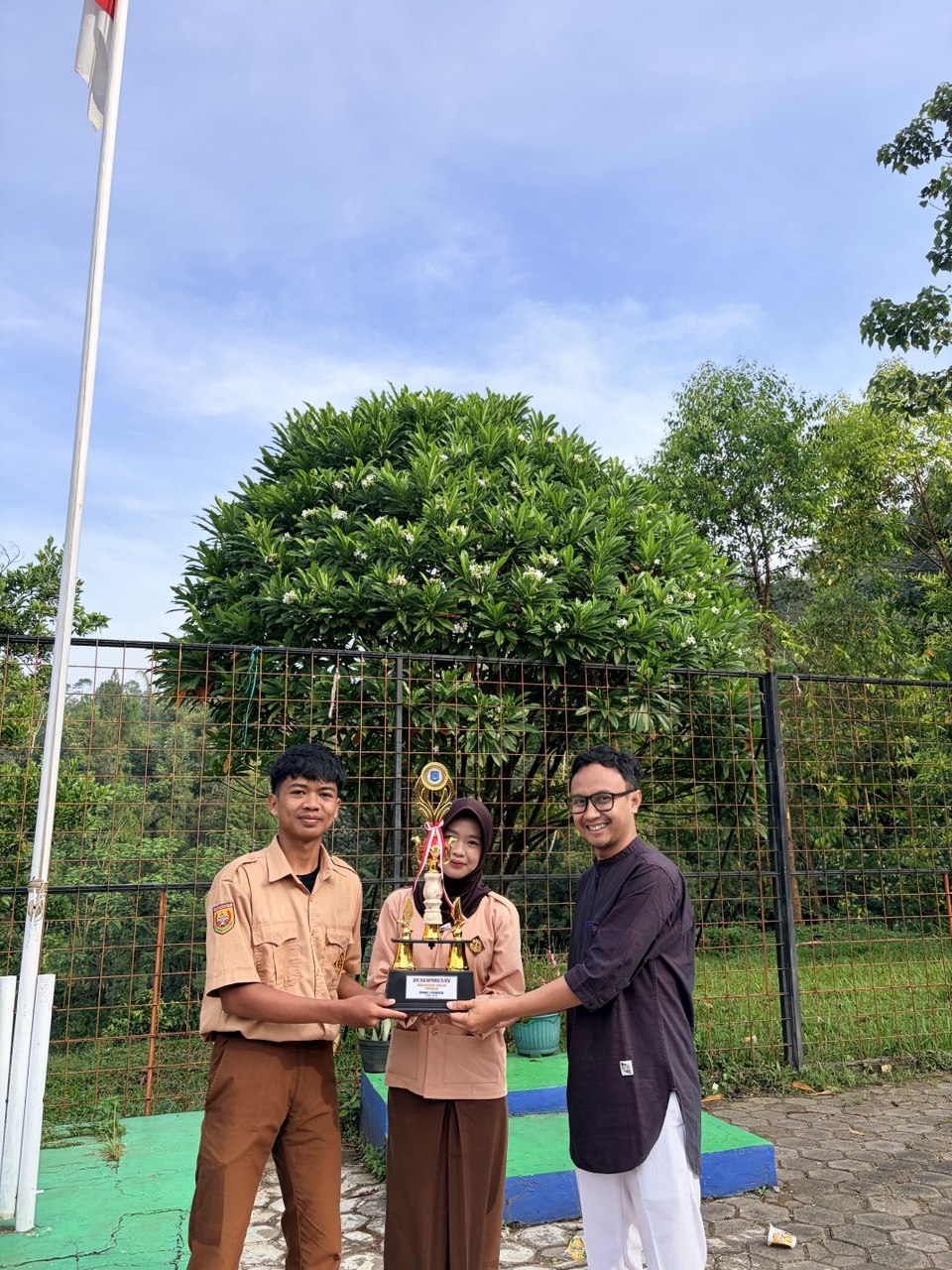 Dua siswa XI RPL 1 memegang piala kehadiran terbaik bersama guru di depan tiang bendera SMKN 1 Rongga